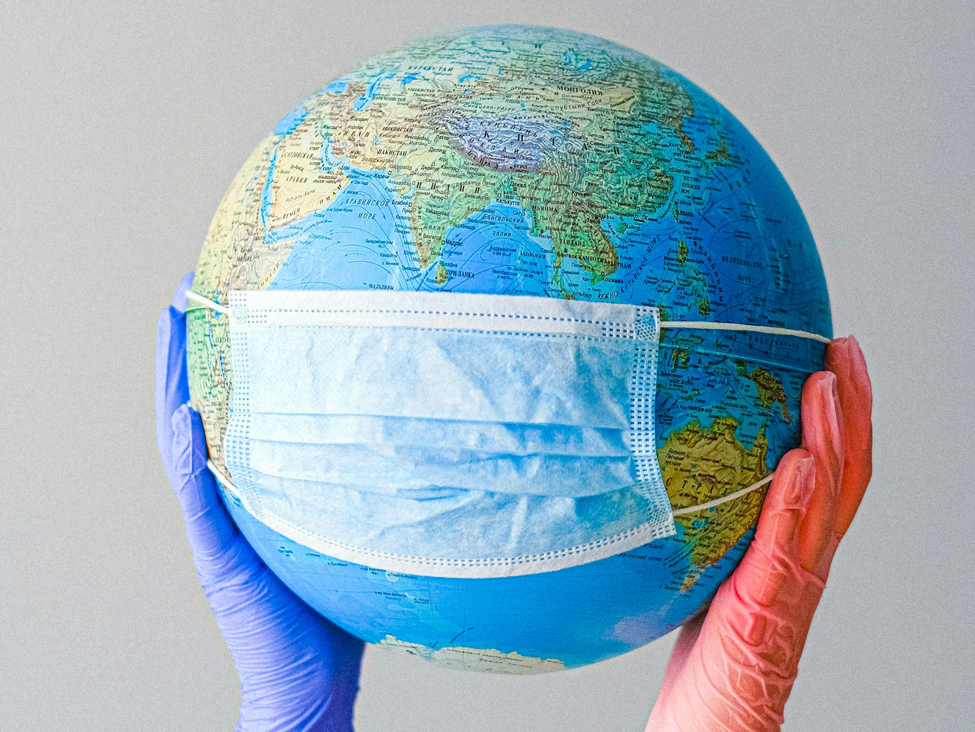 Someone holding up a globe with a surgical mask placed over the front of it to depict the pandemic.