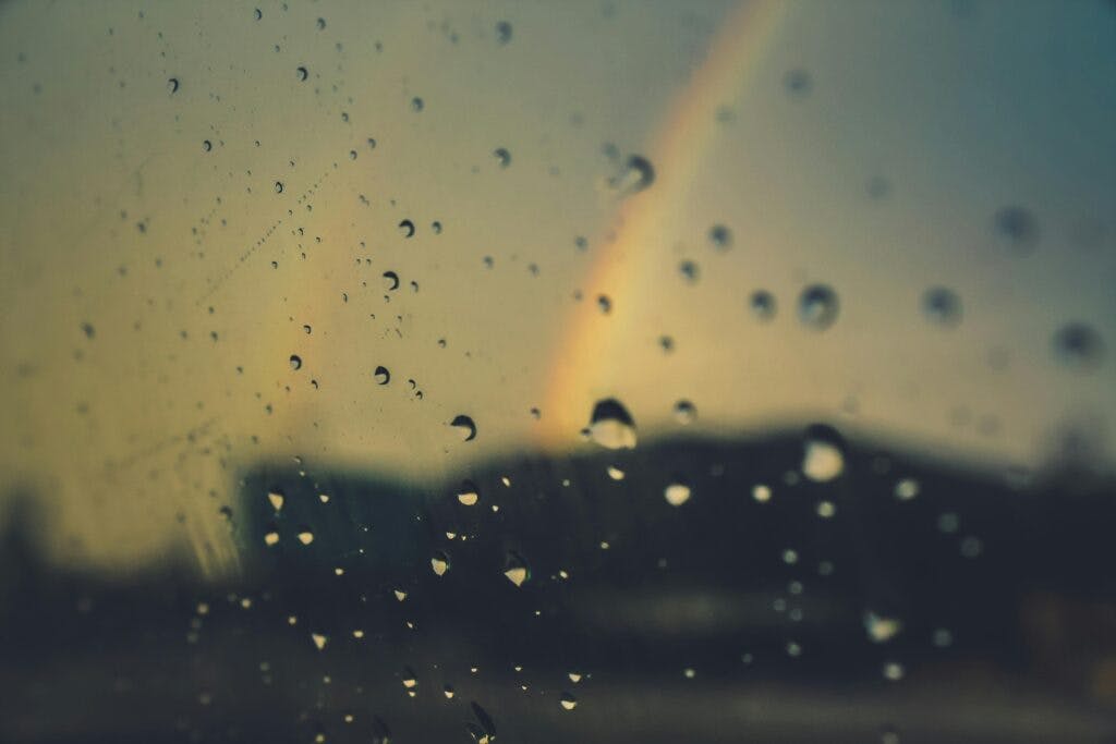 A window with raindrops and a faint rainbow emerging in the background